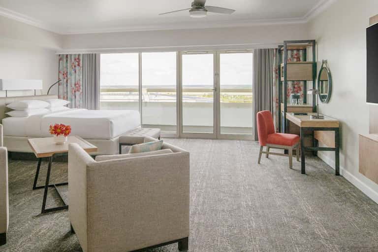 Modern hotel suite with king bed, seating area, and floor-to-ceiling windows overlooking a waterway