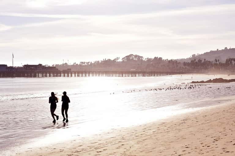 Two runners in wetsuits entering the ocean at a beach with pier and palm trees in background