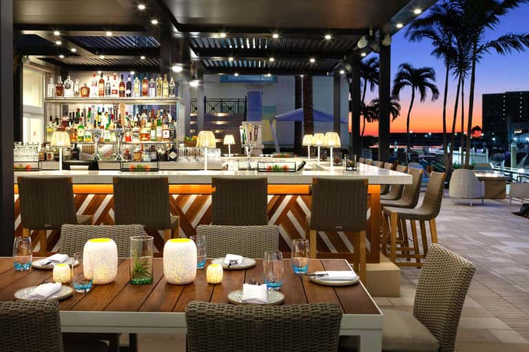 Upscale rooftop bar and dining area at dusk with palm trees, ocean views, and illuminated lounge seating