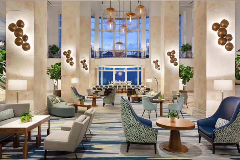 Luxury hotel lobby with high ceilings, marble columns, brass pendant lights, and ocean-view windows