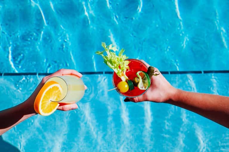 Two hands holding colorful cocktails with citrus garnishes over bright blue pool water