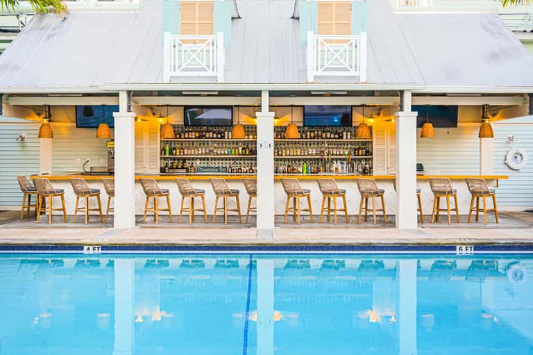 Modern poolside bar with white columns, wooden bar stools, extensive liquor selection, and clear pool water