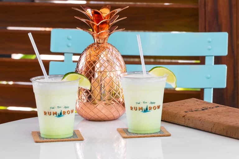 Two tropical cocktails with lime garnish flanking a copper pineapple-shaped drink dispenser on white table