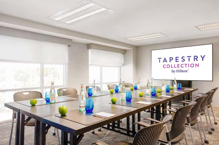 Modern conference room with long table, water bottles, green apples, and Tapestry Collection by Hilton branding on screen