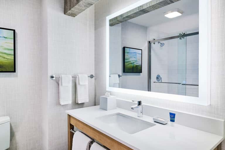 Modern bathroom with illuminated mirror, white vanity, and glass-enclosed shower with landscape artwork