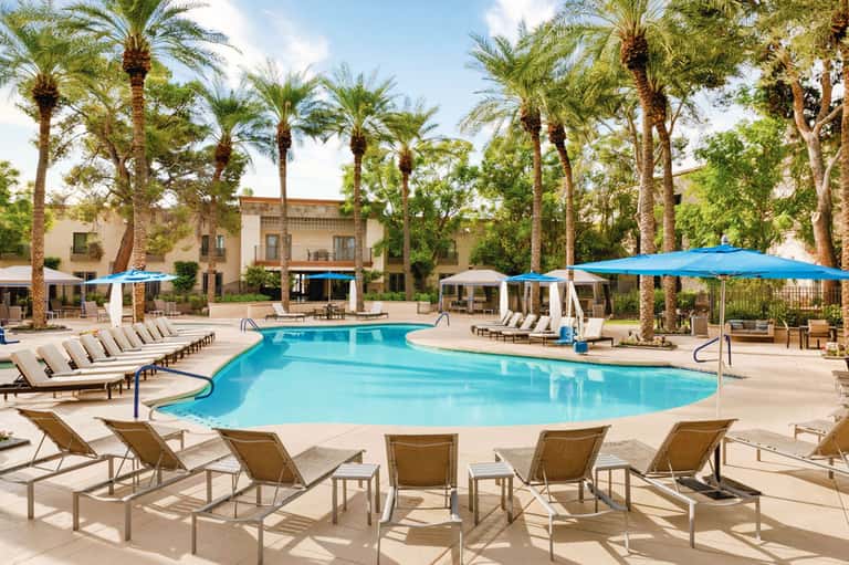 Resort pool with loungers, blue umbrellas, palm trees, and Spanish colonial architecture