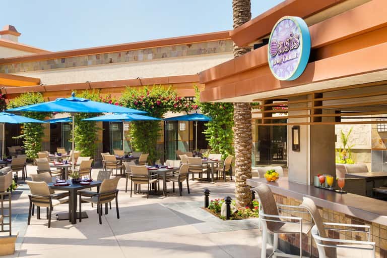 Outdoor restaurant patio with blue umbrellas, tan chairs, and tropical landscaping
