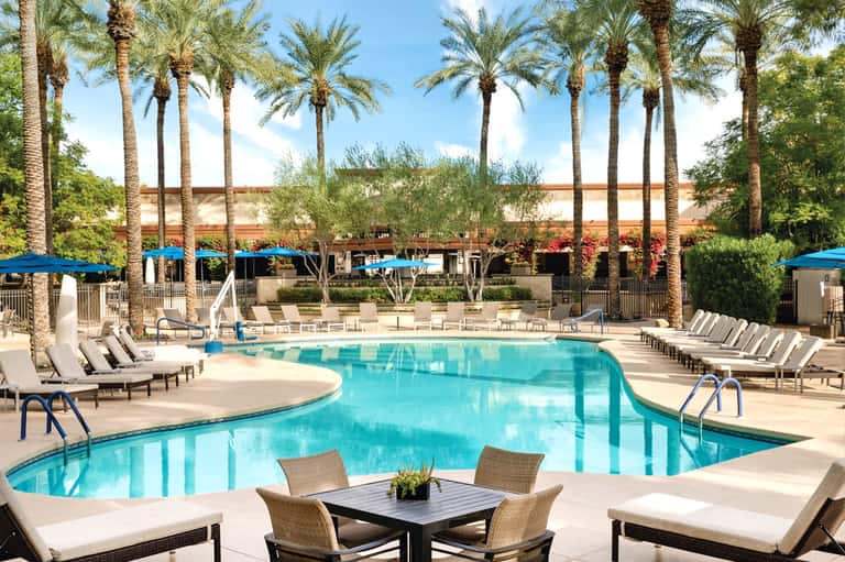 Resort pool surrounded by lounge chairs, blue umbrellas, tall palm trees, and Spanish-style architecture