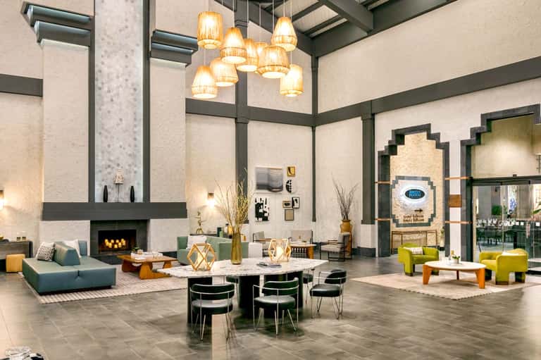 Modern hotel lobby with high ceilings, pendant lights, fireplace, and contemporary furniture in green and yellow tones