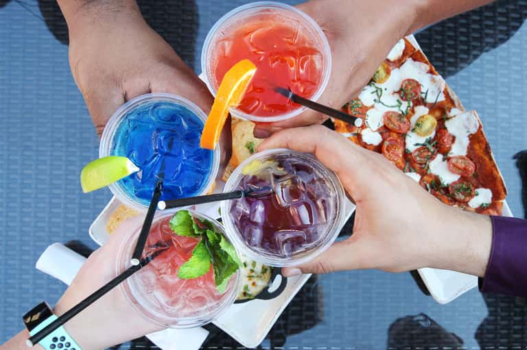 Four colorful cocktails with citrus garnishes and straws held by people's hands at poolside