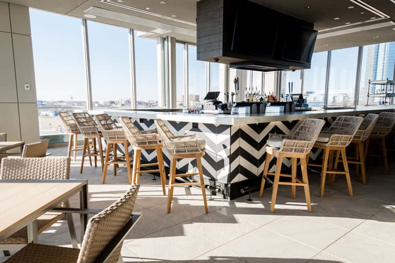 High-rise rooftop bar with chevron-patterned counter, woven bar stools, and expansive city skyline views
