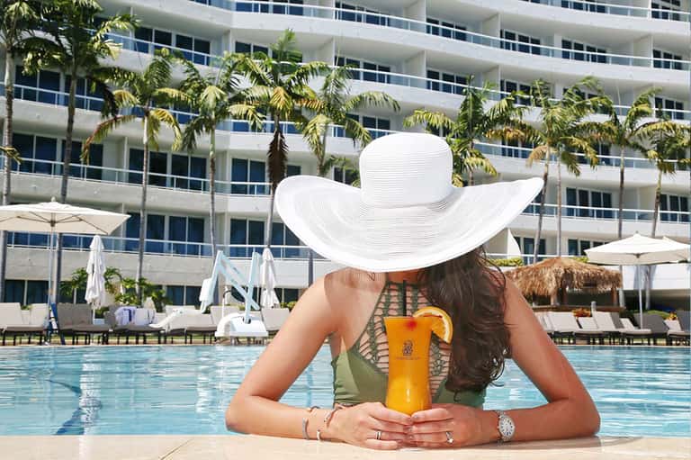 Woman in white hat at resort pool holding tropical drink with high-rise building backdrop