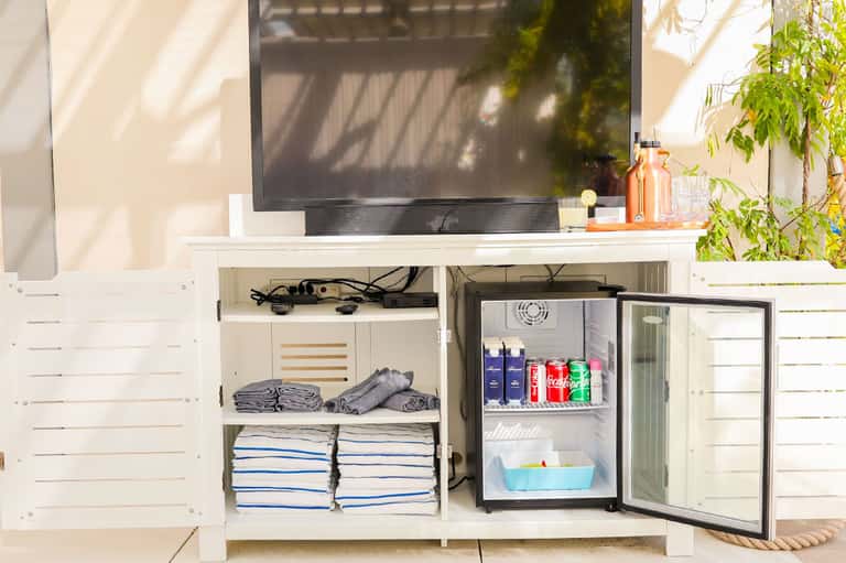White outdoor entertainment cabinet with open door revealing mini refrigerator stocked with beverages and folded towels on shelves