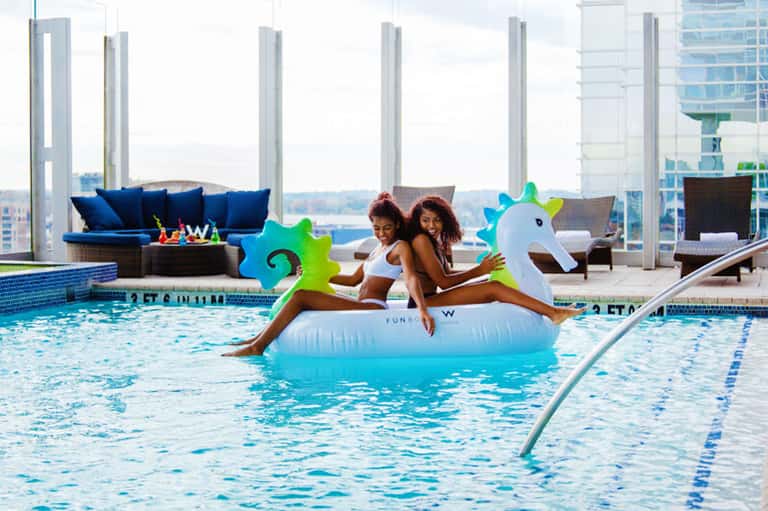 Indoor pool with two women on a unicorn float float against a city skyline backdrop