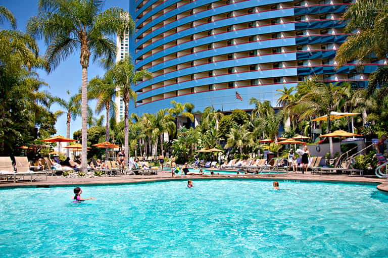 Resort pool with turquoise water, palm trees, blue high-rise hotel building, and guests relaxing on lounge chairs