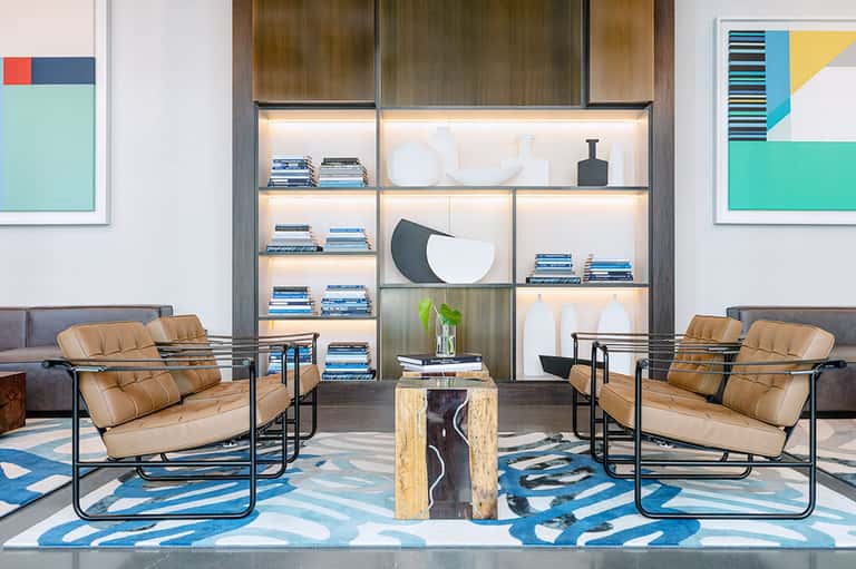 Modern lounge with tan leather chairs, lit shelving unit, and blue patterned rug