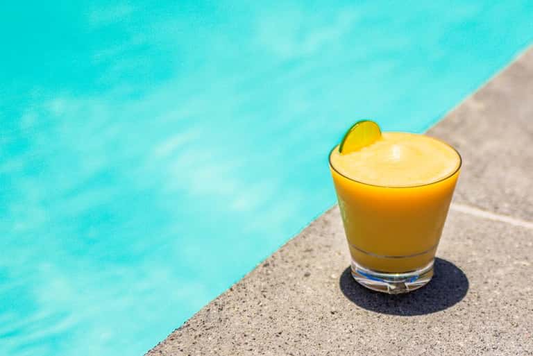 Yellow cocktail with lime slice sitting on pool deck edge next to crystal blue swimming pool