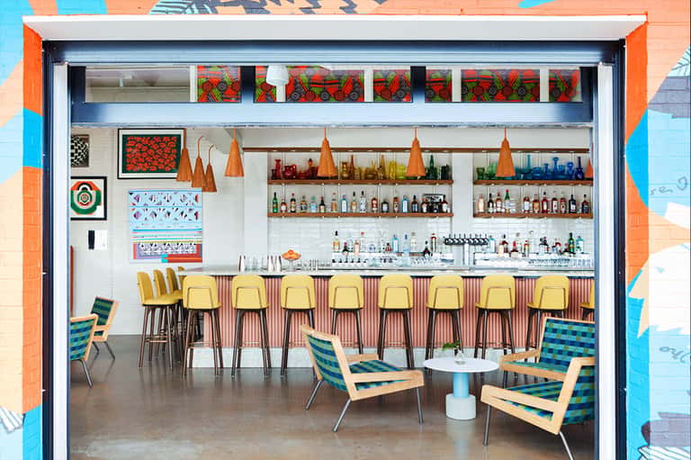 Colorful bar interior with yellow stools, shelves of liquor bottles, and vibrant artwork on exposed brick walls