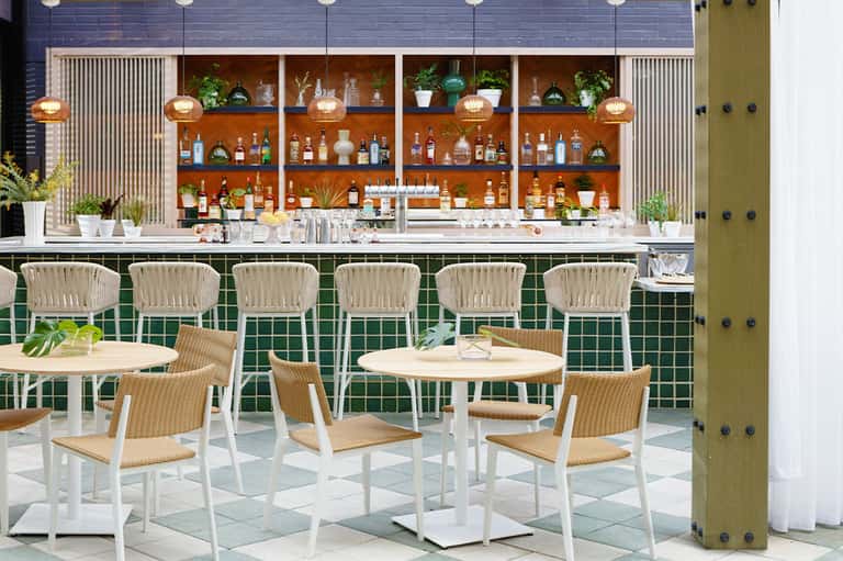 Modern bar lounge with green tile walls, wooden shelving displaying bottles, and woven bar stools