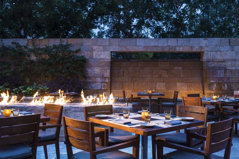 Outdoor dining area with wooden tables and chairs beside fire pits and stone wall at dusk