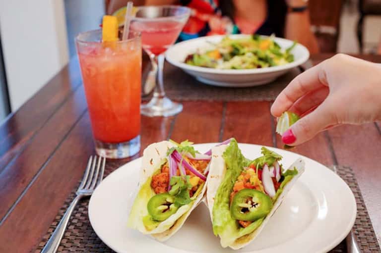 Two fish tacos with lettuce and jalapeños on white plate, colorful cocktails in background at restaurant table