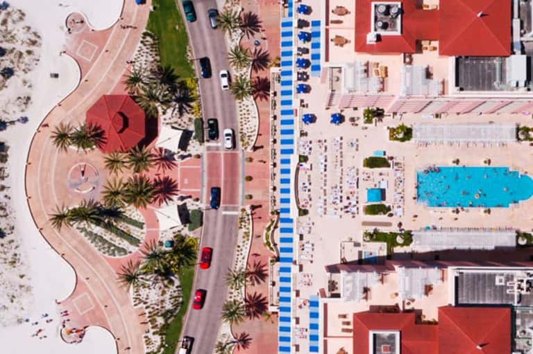 Aerial view of beachfront resort with red-roofed buildings, palm trees, blue umbrellas, and sandy beach