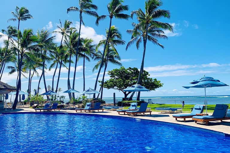Resort pool with palm trees, blue umbrellas, lounge chairs, and ocean view in tropical setting