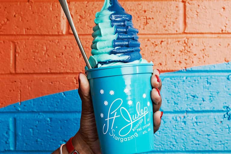 Hand holding turquoise cup with blue and white swirled frozen treat against painted brick wall