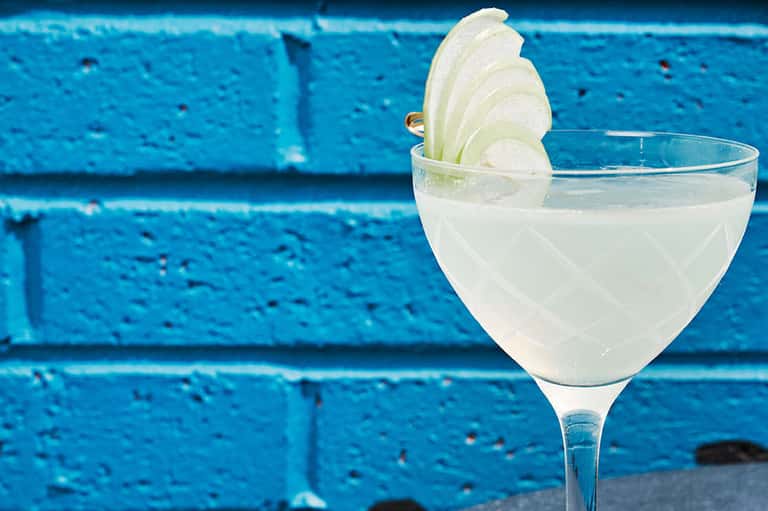 Clear cocktail with cucumber slice garnish against bright blue textured wall background