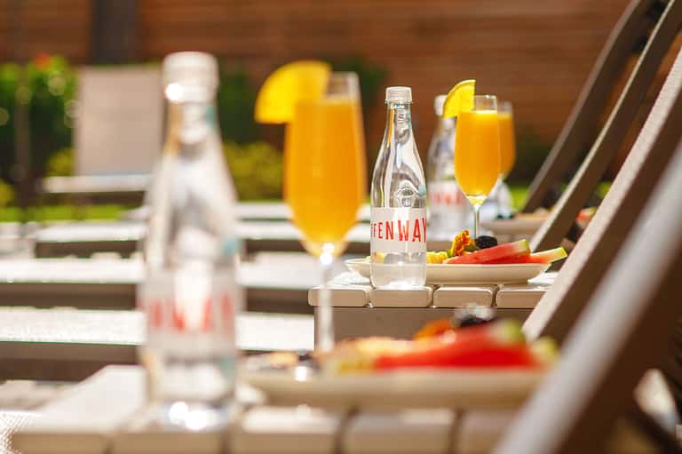 Outdoor rooftop dining setup with orange juice cocktails, fresh vegetables, and water bottles on a sunny terrace.
