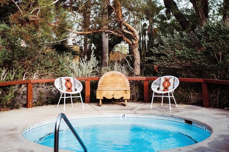 Rustic hot tub surrounded by desert trees with two patterned chairs and wooden bench