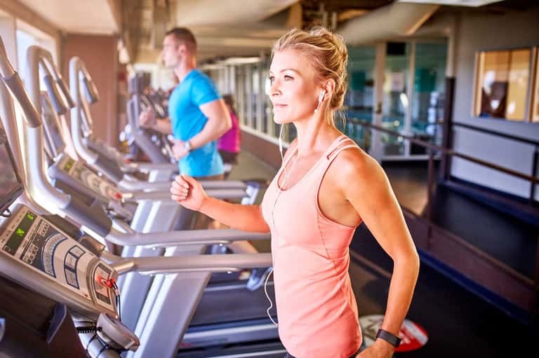 Woman running on treadmill in hotel gym with male guest in background