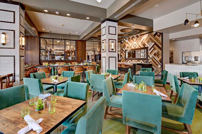 Modern hotel restaurant with turquoise chairs, wooden tables, geometric wall art, and bar area