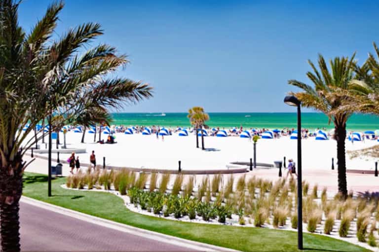 Beachfront resort with white sand, turquoise ocean, palm trees, and manicured coastal landscaping with beach umbrellas