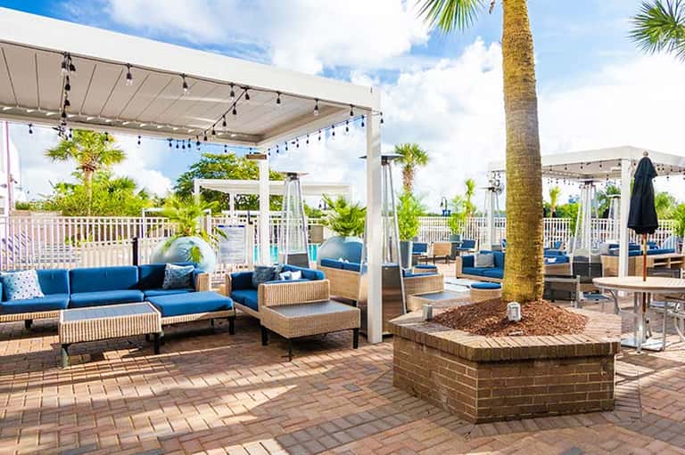 Outdoor resort terrace with blue cushioned seating, woven furniture, white pergolas, palm trees, and ocean views