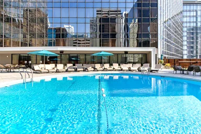 Olympic-sized swimming pool with turquoise water, lounge chairs, umbrellas, and city skyline backdrop