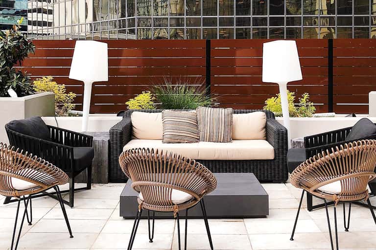 Modern patio with black wicker sofa, rattan chairs, white floor lamps, and wooden fence backdrop with planters