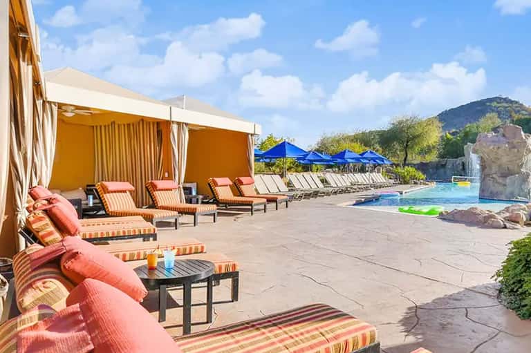Luxurious poolside area with orange cabanas, striped loungers, blue umbrellas, and mountain views