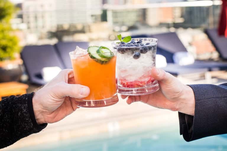 Two people toasting colorful cocktails at a rooftop pool with city backdrop