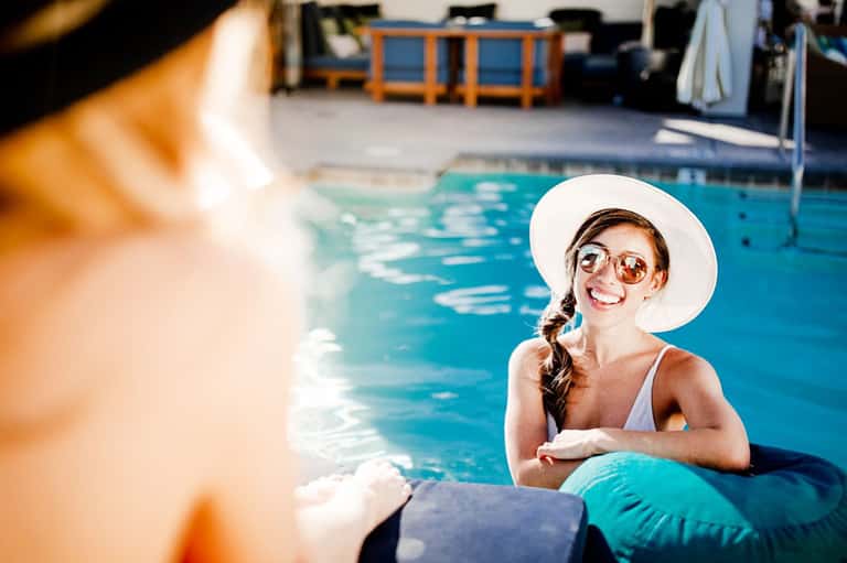 Woman in white sun hat and sunglasses smiling by resort pool edge with lounge chairs