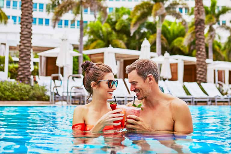 Couple enjoying tropical cocktails in resort pool with palm trees and white cabanas in background