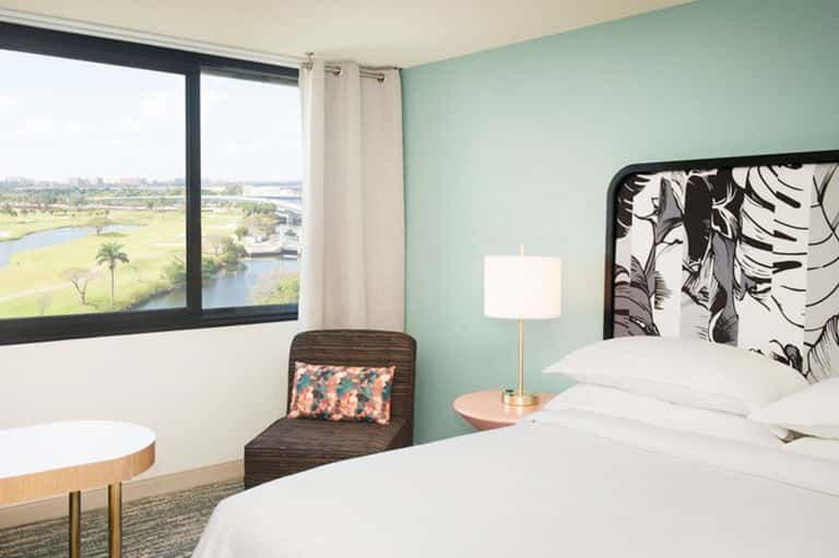 Modern hotel bedroom with mint green walls, white bedding, and large window overlooking scenic landscape