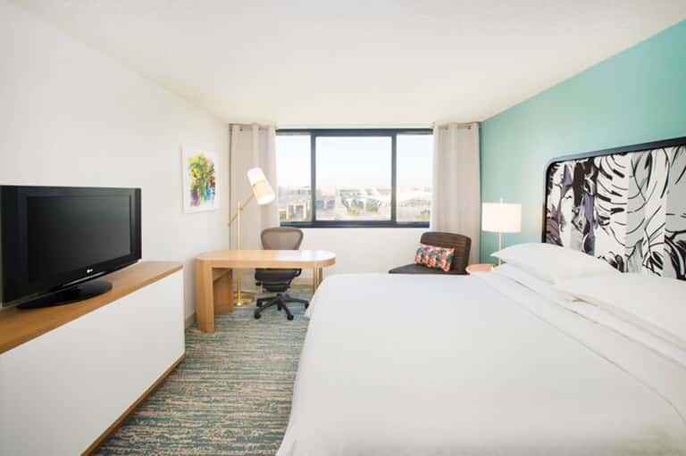 Modern hotel room with turquoise accent wall, queen bed, desk, TV, and city views