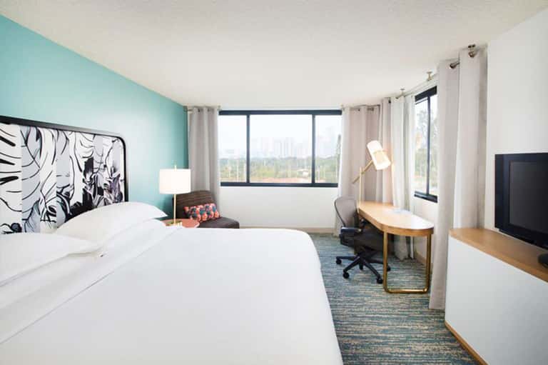 Modern hotel bedroom with turquoise accent wall, two queen beds, desk, and floor-to-ceiling windows with city views