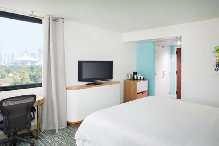 Modern hotel room with white bedding, TV, desk, and city skyline view through window