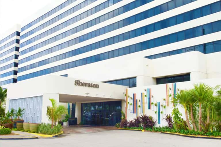 Sheraton hotel entrance with white facade, palm trees, and colorful vertical garden accent
