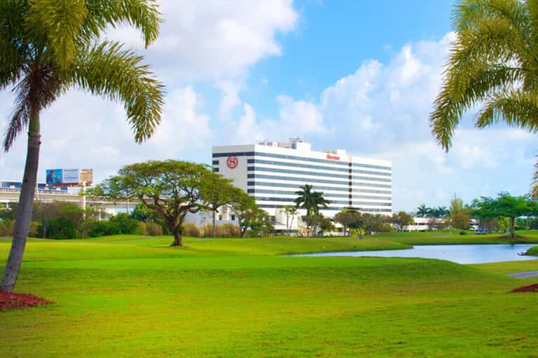 Contemporary hotel building with striped facade beside manicured green lawn, palm trees, and reflecting pond