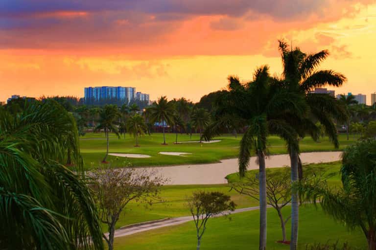 Golf course at sunset with palm trees, water features, and high-rise resort buildings in background