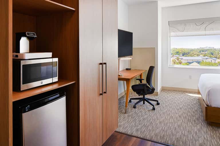 Modern hotel room with wood furnishings, kitchenette, work desk, and window overlooking suburban landscape