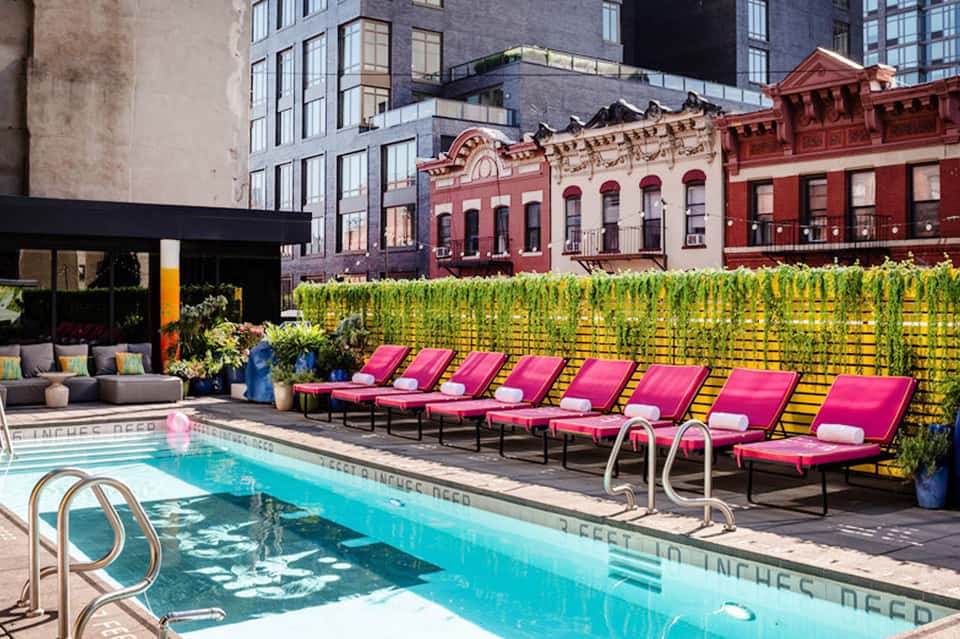 Rooftop pool with bright pink lounge chairs, yellow privacy fence, and urban architecture backdrop